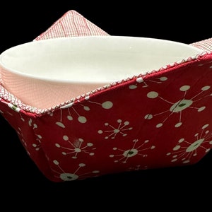 May include: Red fabric bowl cozy with a white and green starburst pattern. The cozy is folded and a white ceramic bowl is inside.
