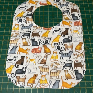 May include: A white fabric baby bib with a cat pattern. The bib has a rounded neckline and a white background with a repeating pattern of colorful cats in various poses.