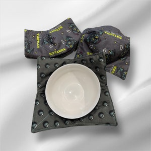 以下が含まれることがあります： ティーンエイジ・ミュータント・ニンジャ・タートルズのテーマのグレーとグリーンのファブリック製鍋つかみ2個セット。鍋つかみには、ティーンエイジ・ミュータント・ニンジャ・タートルズのキャラクターと「Teenage Mutant Ninja Turtles」と「Turtles」の文字が繰り返しパターンで描かれています。鍋つかみは、中央に白いセラミック製のボウルを置いています。