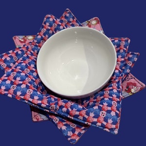 May include: A white ceramic bowl rests on a quilted bowl cosy. The cosy has a blue and pink floral pattern and a scalloped edge. The background is a dark blue.