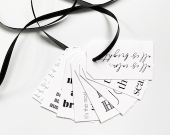 May include: A set of white gift tags with black text. The tags are arranged in a fan shape and have various phrases written on them, including "it's the most", "all is bright", "all is calm", and "the sea". A black ribbon is tied around the top of the tags.