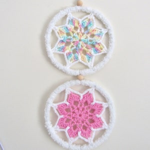 May include: Two white crocheted dreamcatchers with a star-shaped design. The top dreamcatcher has a rainbow coloured star and the bottom dreamcatcher has a pink star.