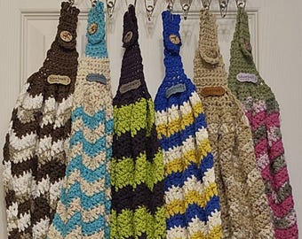 May include: Six crocheted dish towels hanging on a metal rack. The towels are in various colors and patterns, including brown and white, blue and white, green and black, blue and yellow, beige and pink, and green and pink. Each towel has a button and a tag that says "Dishtowel".