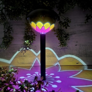May include: A solar powered garden light shaped like a flower with a pink and yellow design. The light casts a pink flower shadow on the ground.