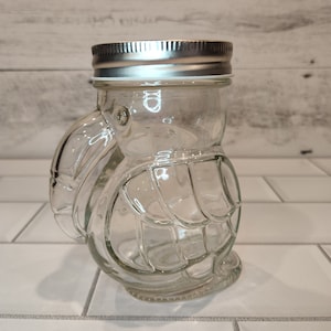May include: Clear glass jar with a silver lid shaped like a turtle. The jar has a textured surface that resembles a turtle shell.