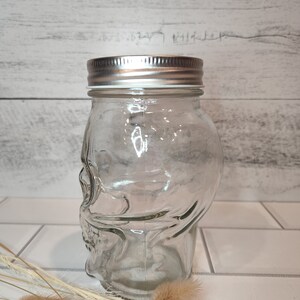 May include: A clear glass jar shaped like a skull with a silver lid. The jar is empty and sitting on a white tiled surface.