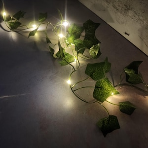 May include: A string of warm white fairy lights wrapped around artificial green ivy leaves. The lights are strung on a thin wire and are spaced evenly apart.
