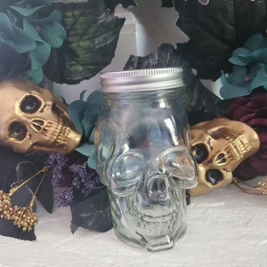 May include: A clear glass jar shaped like a skull with a silver lid. The jar is surrounded by dark green and purple flowers.