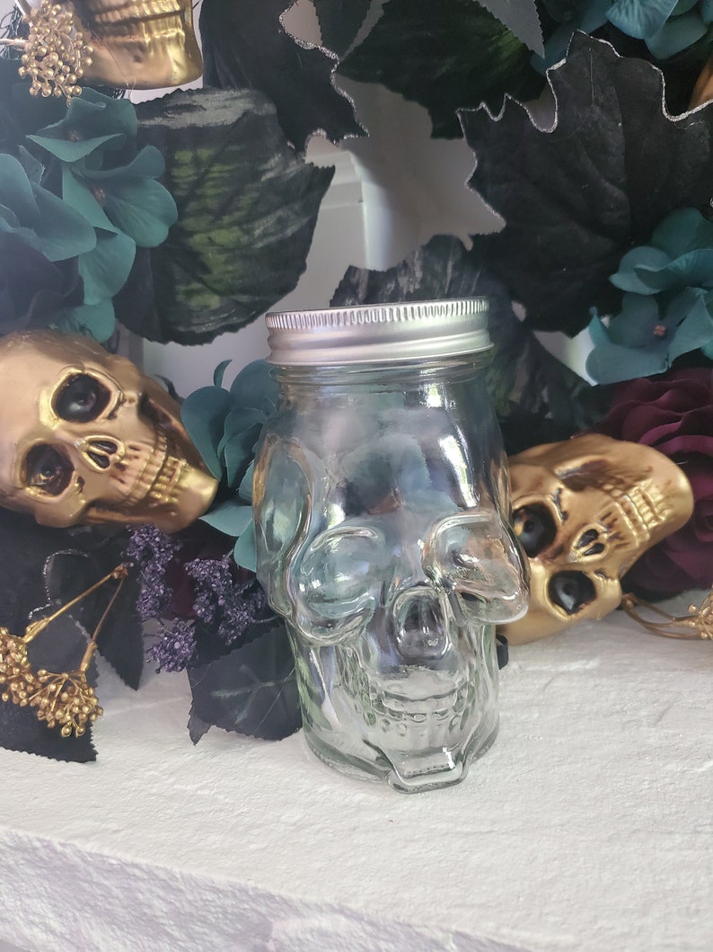 May include: A clear glass jar shaped like a skull with a silver lid. The jar is surrounded by dark green and purple flowers.