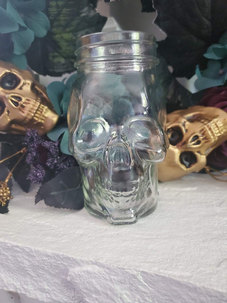 May include: A clear glass jar shaped like a skull. The jar has a textured surface and is sitting on a white surface.