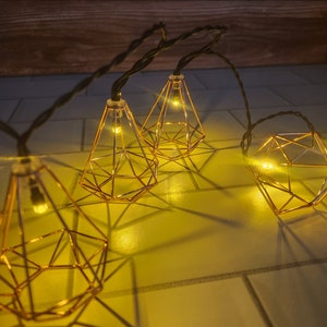 May include: A string of warm white lights with geometric gold cage bulbs. The lights are strung across a white tiled surface.