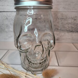 May include: A clear glass jar shaped like a skull with a silver lid. The jar is sitting on a white tiled surface with dried flowers.