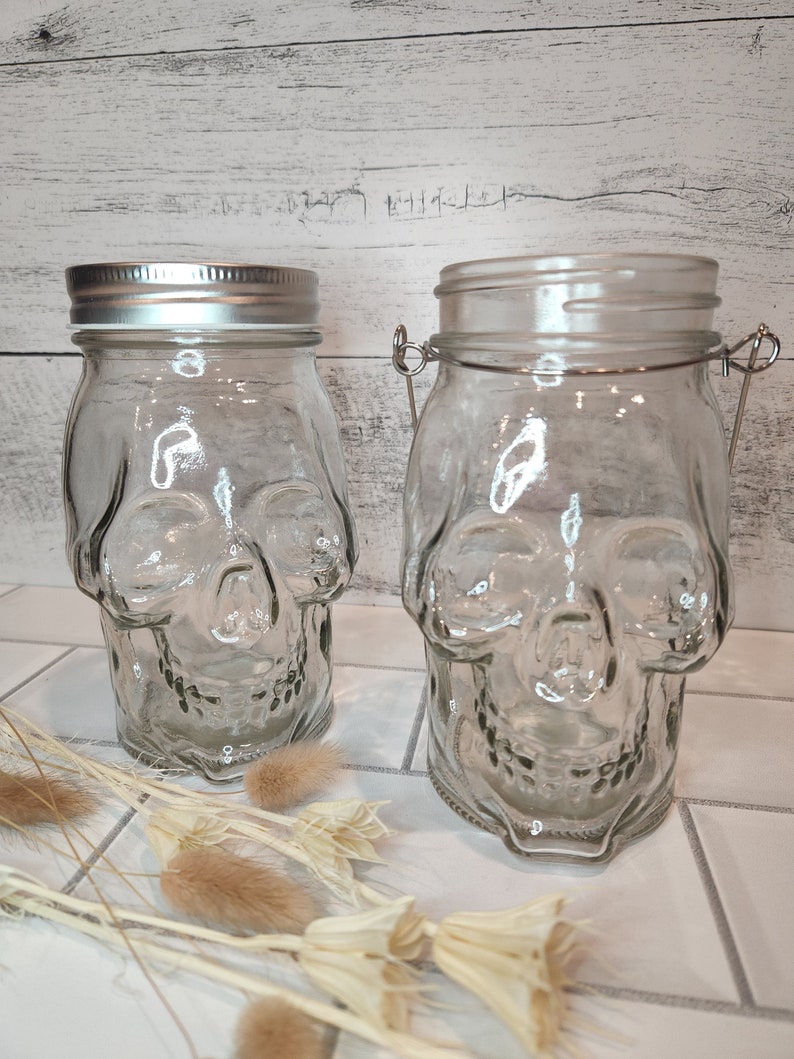 May include: Two clear glass jars shaped like skulls with silver lids. The jars are sitting on a white tiled surface with dried flowers in the foreground.