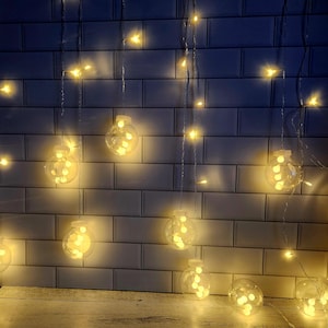 May include: A string of white globe lights with warm white LED bulbs hanging from a white tiled wall. The lights are arranged in a cascading pattern, creating a warm and inviting ambiance.