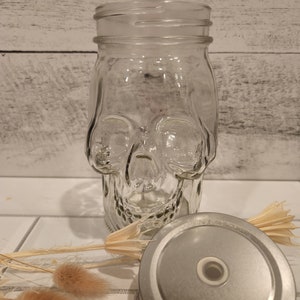 May include: A clear glass jar shaped like a skull with a silver lid. The jar is sitting on a white tiled surface with dried flowers.
