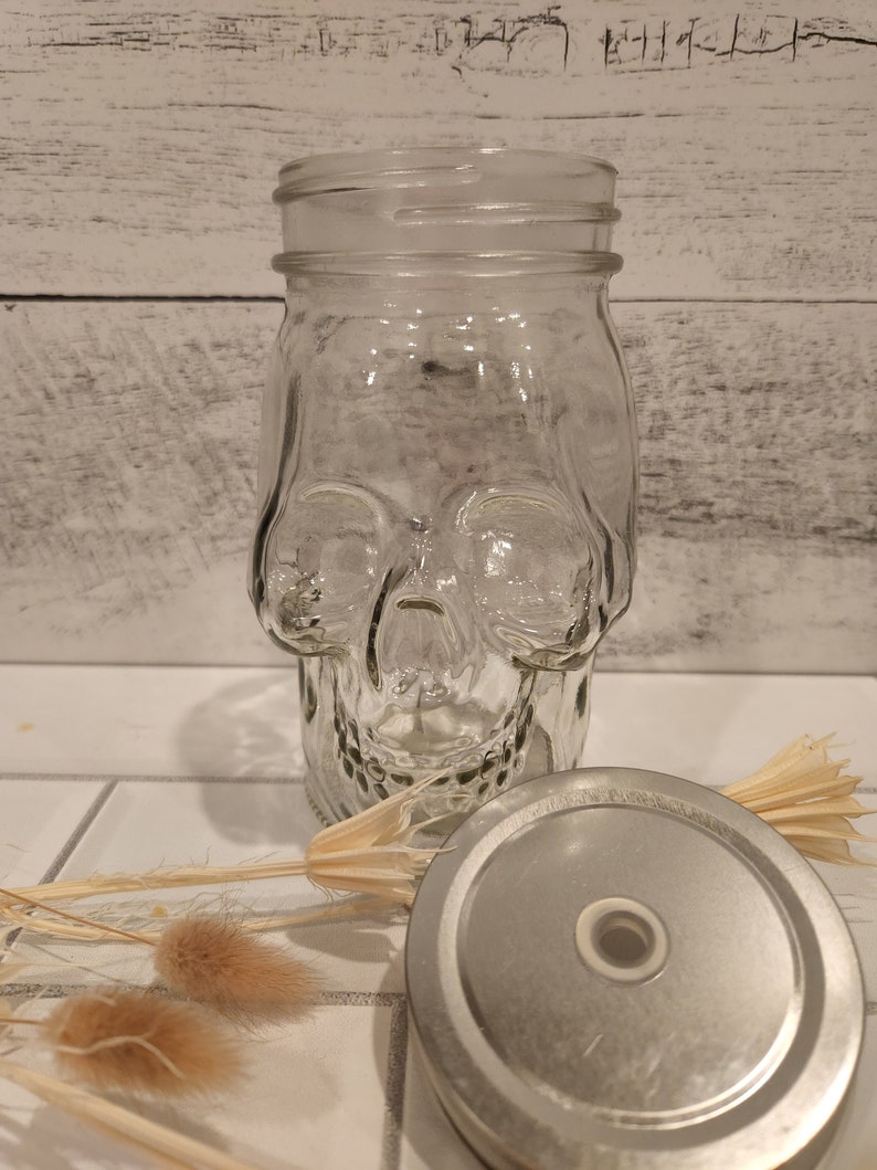 May include: A clear glass jar shaped like a skull with a silver lid. The jar is sitting on a white tiled surface with dried flowers.