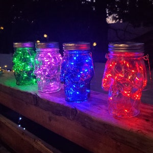 May include: Four glass jars with lids, each containing a string of fairy lights. The jars are lit up in different colors: green, purple, blue, and red.