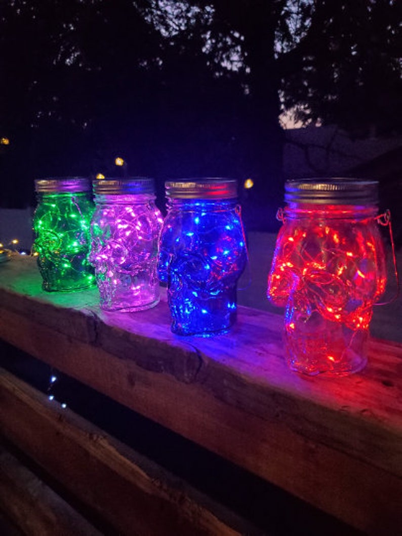 May include: Four glass jars with lids, each containing a string of fairy lights. The jars are lit up in different colors: green, purple, blue, and red.