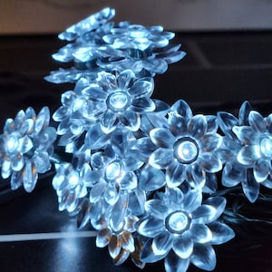 May include: A string of white LED fairy lights with clear plastic flower-shaped bulbs. The lights are strung together with green wire.