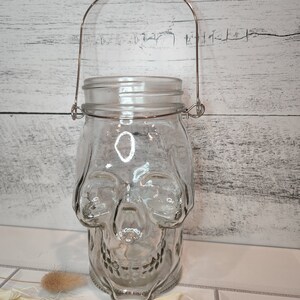 May include: A clear glass jar shaped like a skull with a metal handle. The jar is sitting on a white tiled surface.