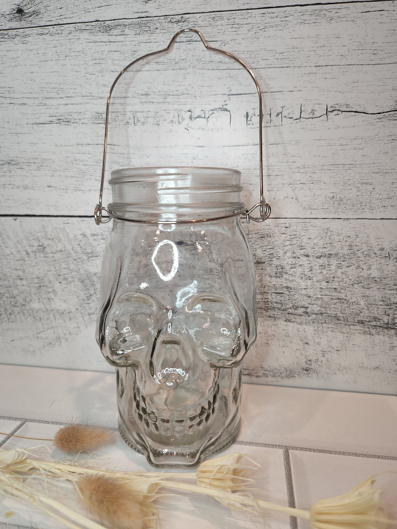 May include: A clear glass jar shaped like a skull with a metal handle. The jar is sitting on a white tiled surface.