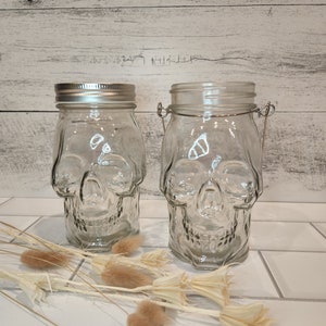 May include: Two clear glass jars shaped like skulls with metal lids. The jars are sitting on a white tiled surface.
