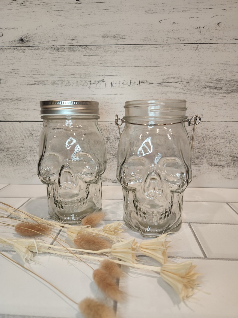 May include: Two clear glass jars shaped like skulls with metal lids. The jars are sitting on a white tiled surface.
