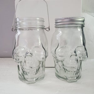 May include: Two clear glass jars shaped like skulls. One jar has a metal handle. Both jars have silver lids.