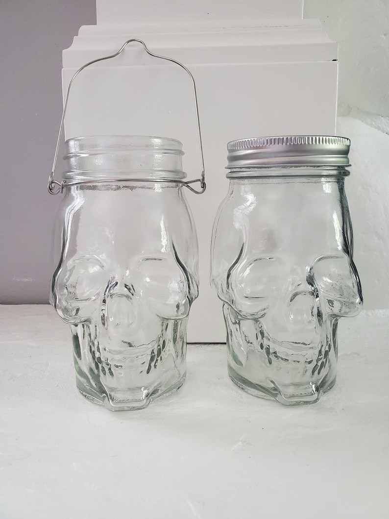 May include: Two clear glass jars shaped like skulls. One jar has a metal handle. Both jars have silver lids.