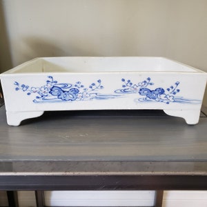 May include: A white ceramic planter with blue and pink floral designs. The planter has four short legs and is rectangular in shape.
