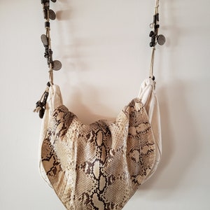 May include: A cream-colored shoulder bag with a snakeskin pattern on the front. The bag hangs from a decorative rope with metal accents and small, round, metal embellishments. The bag has a soft, draped design.