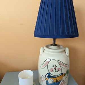 May include: A white ceramic lamp base with a hand-painted design of a bunny holding a carrot. The lamp has a blue pleated lampshade.