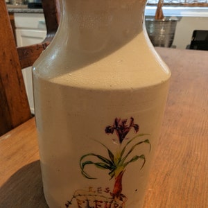 May include: A white ceramic vase with a floral design. The vase has a wide mouth and a narrow base. The floral design features a purple iris with green leaves and the text "LES FLEURS".