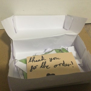 Può includere: Una scatola di cartone bianca con un biglietto di ringraziamento scritto a mano che dice "thank you for the order!" all'interno. La scatola è aperta e c'è carta velina bianca all'interno.
