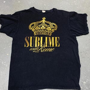 May include: Black t-shirt featuring a gold crown design above the words "SUBLIME with Rome". The shirt is made of a soft material and has a crew neck. The design is centered on the front of the shirt.