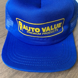 May include: A blue trucker hat with a yellow brim button and mesh back. The front of the hat features a yellow and black logo that reads "AUTO VALUE PREMIUM PRODUCTS". The hat is made of a durable fabric.