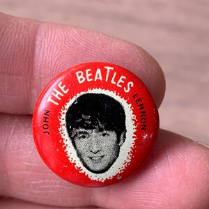 May include: A red button featuring a black and white image of John Lennon, with the text "THE BEATLES" and "JOHN LENNON" in white lettering. The button is round and has a white border around the image.