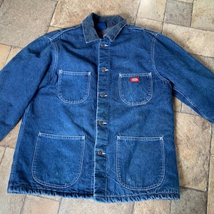 May include: A blue denim chore coat featuring a dark corduroy collar, long sleeves, and a button-front closure. The jacket includes two chest pockets and two large lower pockets, with a small red Dickies brand label on the upper left pocket.