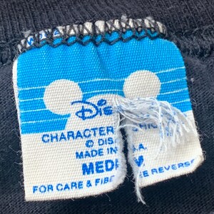 May include: Close-up of a black t-shirt label with a blue and white design. The label features the Disney logo and the words "CHARACTER" and "MADE IN". The size is "MEDIUM".