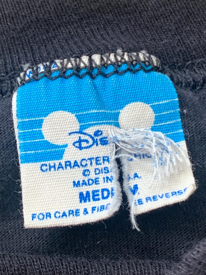 May include: Close-up of a black t-shirt label with a blue and white design. The label features the Disney logo and the words "CHARACTER" and "MADE IN". The size is "MEDIUM".