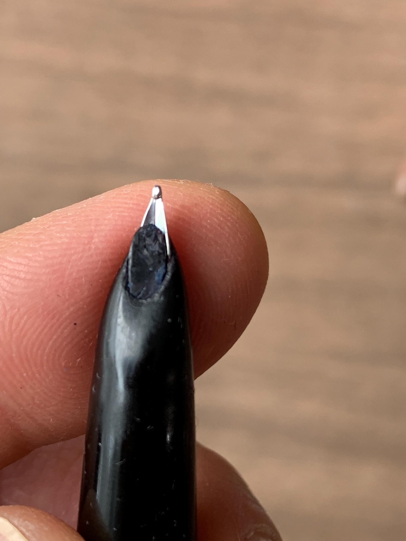 Pu&ograve; includere: Primo piano di un pennino di penna stilografica nero. Il pennino &egrave; argentato e presenta una piccola fessura. Il corpo della penna &egrave; nero e lucido. Lo sfondo &egrave; sfocato e di tono neutro.