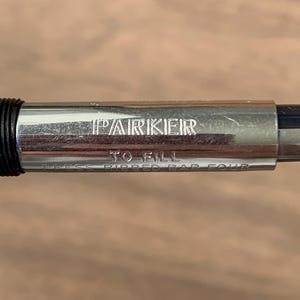 Pu&ograve; includere: Primo piano del corpo argentato di una penna Parker con il nome del marchio inciso. &Egrave; visibile anche il testo "TO FILL" e "PRESS RIBBED BAR FOUR". La penna ha una sezione di presa nera e uno sfondo sfocato.