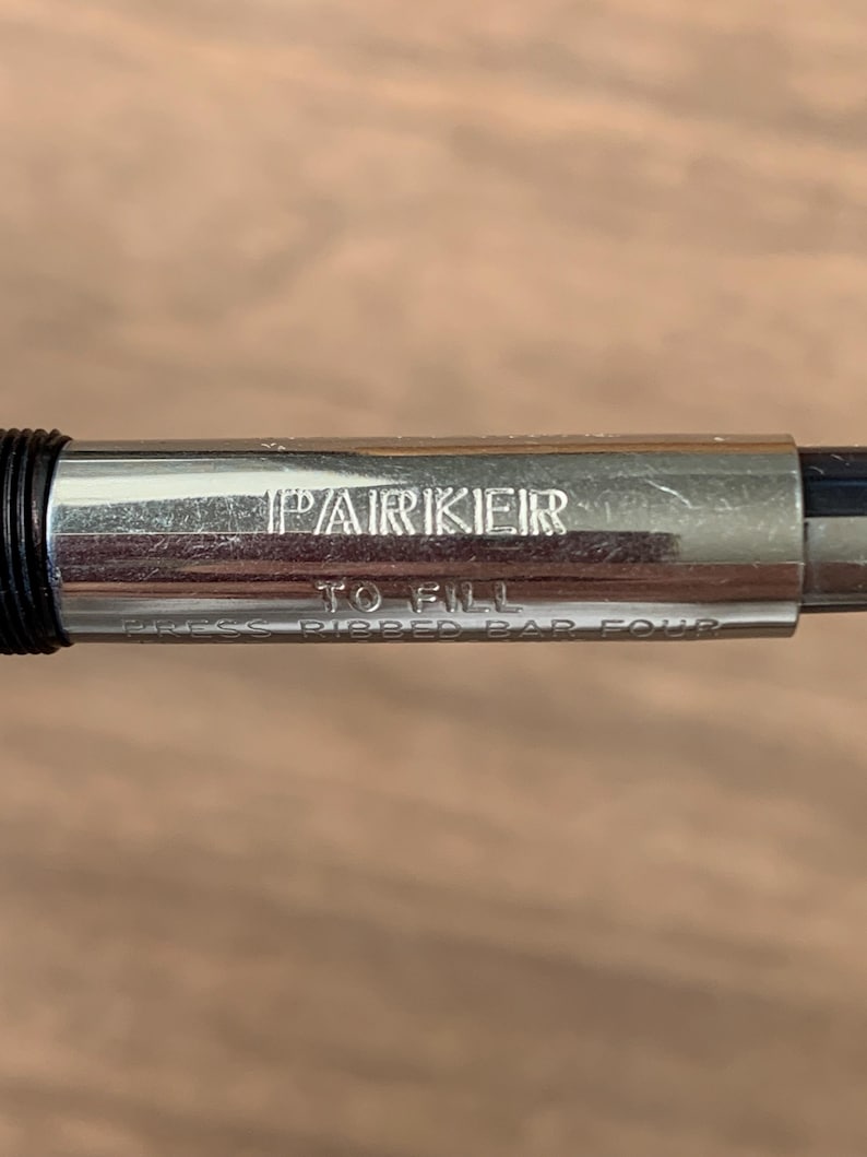 Pu&ograve; includere: Primo piano del corpo argentato di una penna Parker con il nome del marchio inciso. &Egrave; visibile anche il testo "TO FILL" e "PRESS RIBBED BAR FOUR". La penna ha una sezione di presa nera e uno sfondo sfocato.