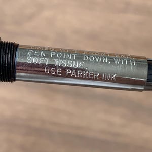 Pu&ograve; includere: Primo piano di un pennino d'argento con dettagli neri. La penna ha un testo inciso che recita: "TIMES, WIPE FRONT END, PEN POINT DOWN, WITH SOFT TISSUE. USE PARKER INK."