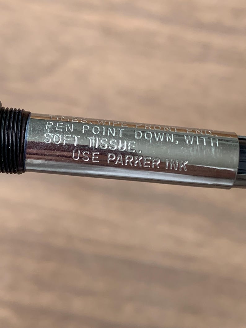 Pu&ograve; includere: Primo piano di un pennino d'argento con dettagli neri. La penna ha un testo inciso che recita: "TIMES, WIPE FRONT END, PEN POINT DOWN, WITH SOFT TISSUE. USE PARKER INK."