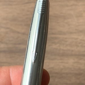 Pu&ograve; includere: Primo piano di una penna color argento con la scritta "PARKER" visibile. La penna ha una finitura metallica lucida e una parte superiore strutturata. La penna &egrave; tenuta su uno sfondo sfocato di legno.
