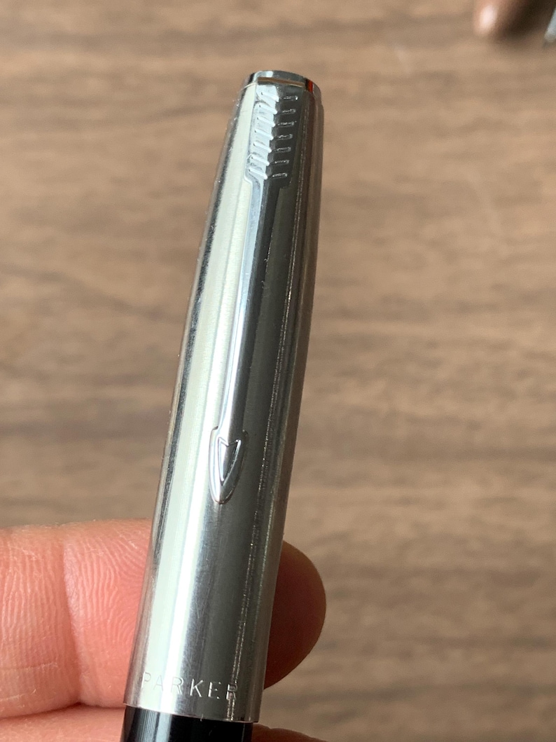 Pu&ograve; includere: Primo piano di una penna color argento con la scritta "PARKER" visibile. La penna ha una finitura metallica lucida e una parte superiore strutturata. La penna &egrave; tenuta su uno sfondo sfocato di legno.