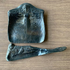 May include: Two antique metal ashtrays. One is a rectangular tray with a woman figure in the center. The other is a long, narrow ashtray with a woman figure on one end. Both are dark gray and sit on a wooden surface.