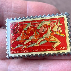 May include: A rectangular pin resembling a postage stamp. The pin features a red background with stylized figures in orange, green, and tan. Text includes "22 USA" and "Pan-American Games Indianapolis 1987". The pin has a silver border.