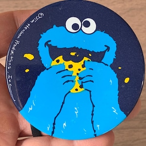May include: A round, navy blue button featuring a cartoon Cookie Monster character eating a cookie. The character is blue with white eyes and is holding a yellow cookie with brown chips. The text "© Jim Henson Productions, Inc." is printed on the button.